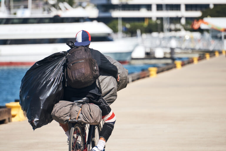 Homeless,Man,Riding,His,Bicycle,While,Carrying,Bags,And,Backpacks.